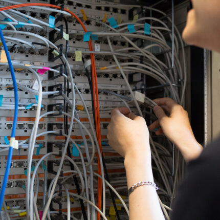 Farbfoto: Serverschrank mit bunten Kabeln und seitlcih Arme einer stehenden Person, die Kabel steckt